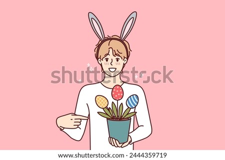 Teenage guy celebrating easter holds pot of plants in shape of multi-colored eggs, wears bunny ears on head. Boy invites to celebrate easter together, or buy decorations to create festive atmosphere