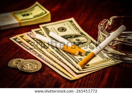 Two cigarettes are on the dollars, lies next to a pack of bills and other cigarette on the glass ashtray. Focus on the cigarettes, image vignetting