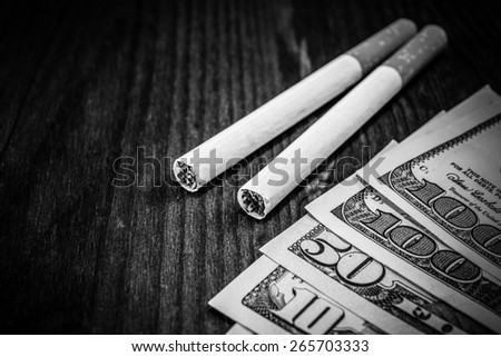 Couple of cigarettes with a several dollar bills on the table. Focus on the cigarettes, image vignetting and black and white tones
