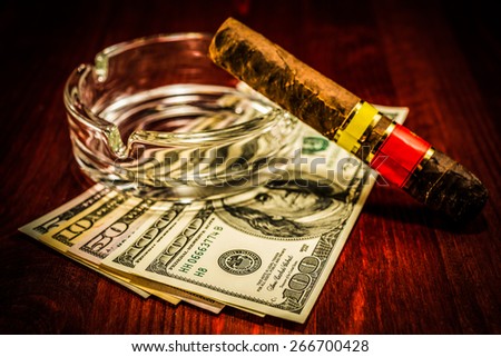 Cuban cigar with glass ashtray on a several dollar bills on the table. Focus on the dollars, image vignetting