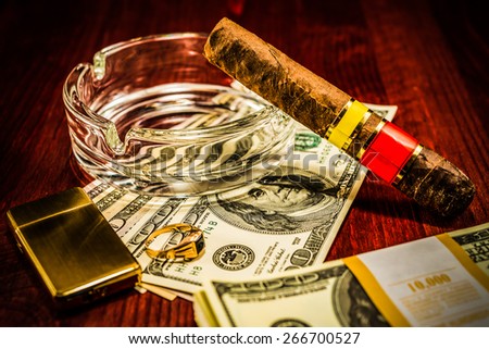 Cuban cigar with glass ashtray on a several dollar bills and also pack of dollars and golden lighter and ring on the table. Image vignetting