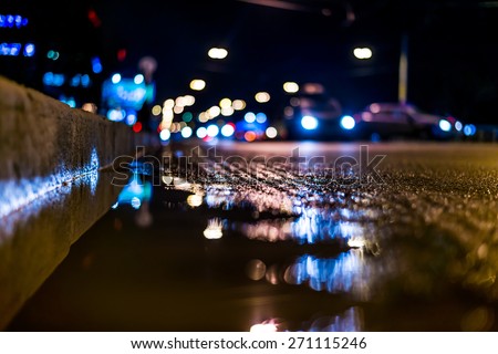 Night city after rain, a reflection of the city at night in the water. View of the driving cars from the roadside at the asphalt level, in blue tones