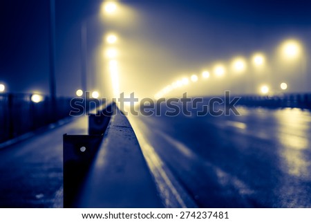 The bright lights of the city at night, the headlights of the approaching cars on the road bridge. View of the highway with a dividing border, image in the yellow-blue toning