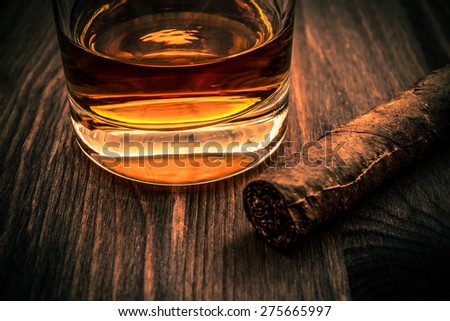 Glass of whiskey and cuban cigar on a wooden table. Focus on the glass of whiskey, image vignetting and the orange-blue toning
