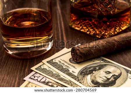 Decanter of whiskey and a glass with cuban cigar and money on a wooden table. Angle view, focus on the cuban cigar
