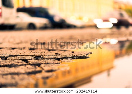 City central square paved with stone after a rain, headlights from car. View from the pavement level next to the roadside puddle, image in the yellow toning