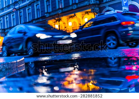 The bright lights of the evening city, driving cars. View from the pavement level next to the roadside puddle, image in blue tones