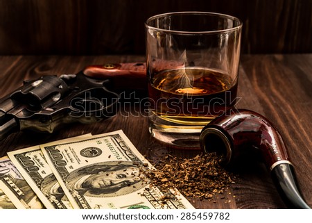 Glass of whiskey and revolver with a money and tobacco pipe on the wooden table. Angle view, focus on the tobacco pipe