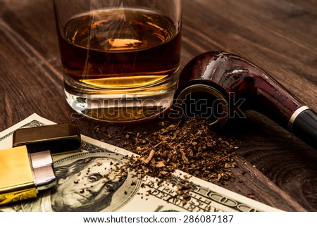 Glass of whiskey with a money and tobacco pipe with tobacco leaves are scattered on the wooden table. Focus on the tobacco pipe