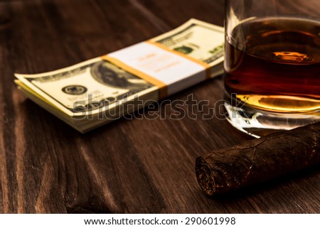 Glass of whiskey and a pack of dollars with cuban cigar on a wooden table. Close up view, focus on the cuban cigar