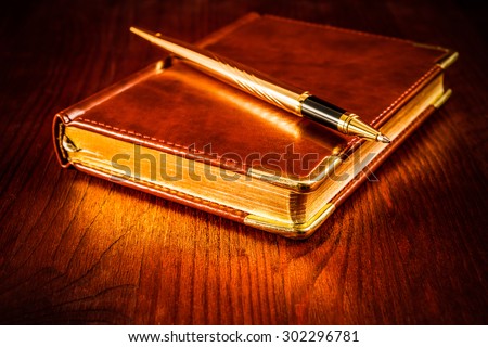 Golden pen on a leather diary on a mahogany table. Image vignetting and hard tones