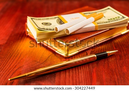 Pack of dollars on a leather diary and cigarettes with golden pen on a mahogany table. Focus on the cigarettes