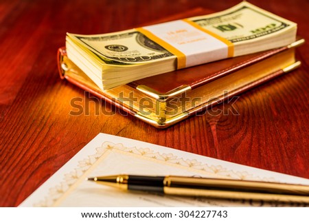 Pack of dollars on the leather diary with bank check and golden pen on the table. Focus on the leather diary
