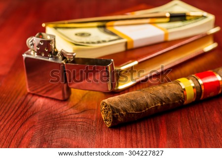 Pack of dollars and open lighter with a leather diary and cuban cigar on a mahogany table. Focus on the cuban cigar