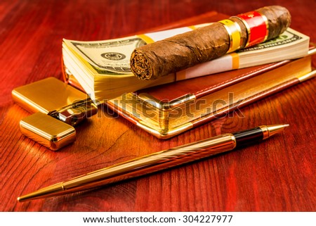 Pack of dollars and open golden lighter with a leather diary and cuban cigar on a mahogany table. Focus on the cuban cigar
