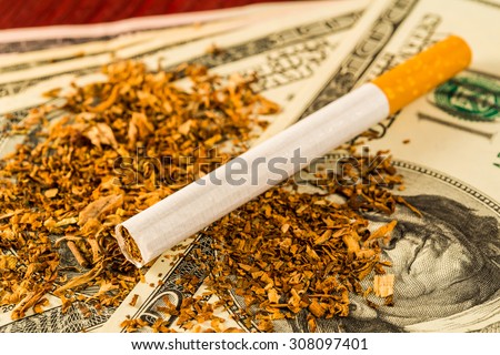 Cigarette and tobacco leaves lie scattered on the dollar bills on a wooden table. Close up view