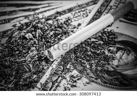 Cigarette and tobacco leaves lie scattered on the dollar bills on a wooden table. Close up view, image vignetting in hard black and white tones