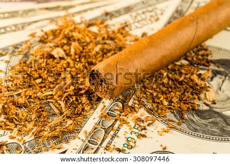 Cuban cigar and tobacco leaves lie scattered on the dollar bills on a wooden table. Close up view