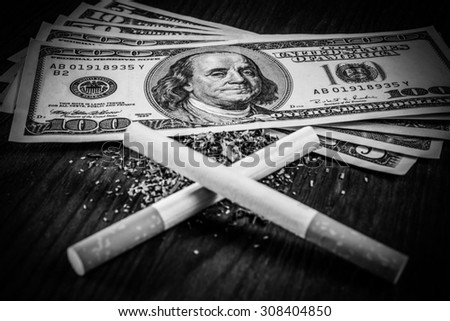 Two cigarettes lying crosswise on a fill of tobacco and dollar bills on the table. Close up view, focus on the dollars, image vignetting in hard black and white tones