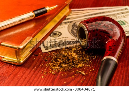 Tube for smoking tobacco and money with leather diary and golden pen on a wooden table. Close up view, focus on the tube