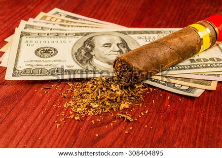 Cuban cigar and a fill of tobacco with the dollar bills on a wooden table. Close up view, focus on the cuban cigar