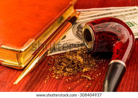 Tube for smoking tobacco and money with leather diary and golden pen on a wooden table. Close up view, focus on the tube
