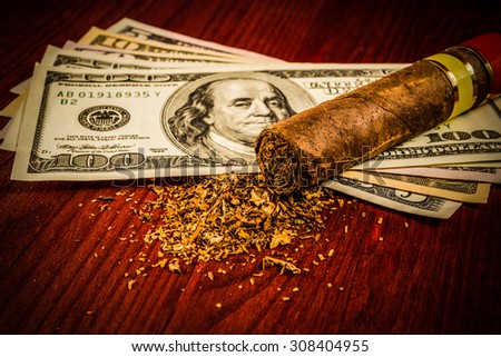 Cuban cigar and a fill of tobacco with the dollar bills on a wooden table. Close up view, focus on the cuban cigar, image vignetting and hard tones