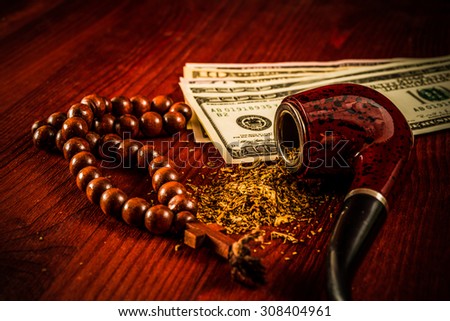 Tube for smoking tobacco with money and rosary with a cross on a wooden table. Close up view, focus on the tube, image vignetting and hard tones
