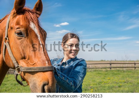 Happy beautiful brunette taking care of her domestic horse outdoors. Selective focus on the horse, narrow depth of field, direct sunlight, darker shadows