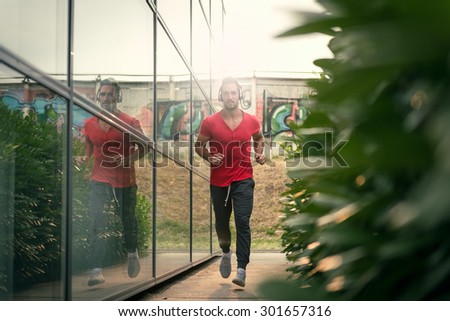 Young male jogging next to modern sport center, listening music with his headphones on. Wearing shirt and tracksuits. Selective focus.