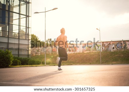 Young male jogging next to modern sport center, listening music with his headphones on. Wearing tracksuits without shirt. Toned image, direct sunlight, lens flare.