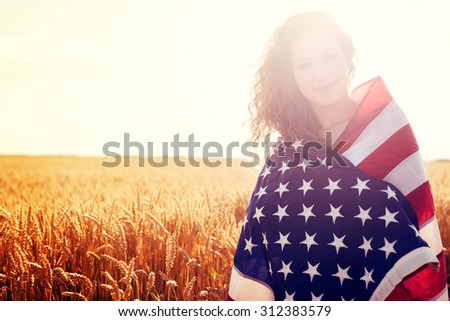 Young smiling girl covered with USA flag in golden wheat field.