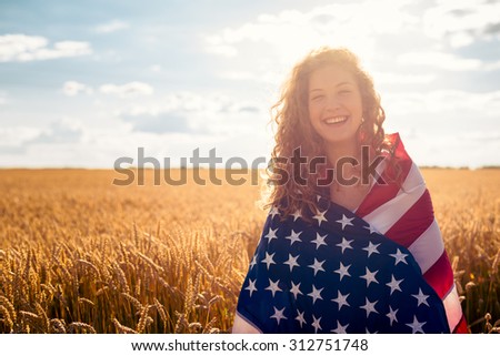 Young smiling girl covered with USA flag in golden wheat field. Selective focus. Lens flare.