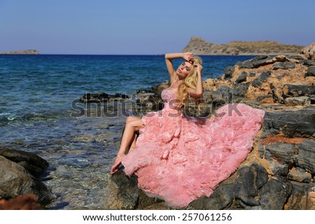 Beautiful blond woman with long legs lying on the background of a beautiful view of the sea in a fantastic pink dress