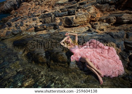 Beautiful blond woman with long legs lying on the background of a beautiful view of the sea in a fantastic pink dress