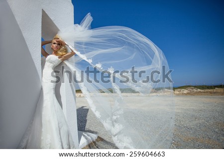A young beautiful blond bride stands on a fabulous balcony with beautiful wooden roof in a luxury hotel in a white wedding dress unique in Santorini on the background of the sunny sky