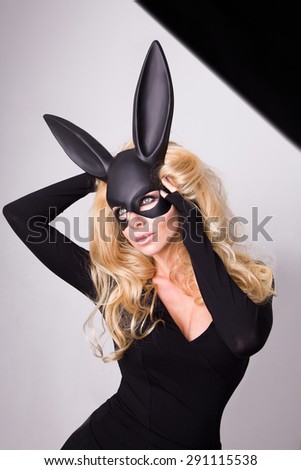 Beautiful blonde-haired young woman in carnival mask ballroom rabbit with long ears sensual sexy in a black dress, standing defiantly on a white background.