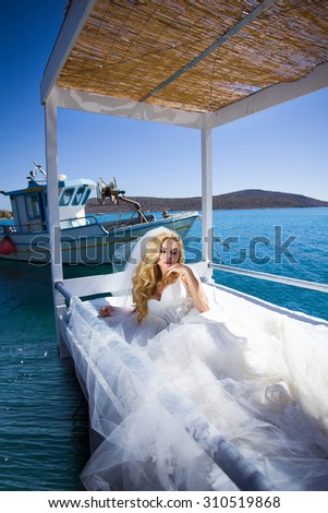 Beautiful blond woman with long legs in a white ball gown wedding dress it is lying on the bed beach  on the water