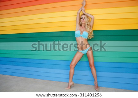 Very beautiful blonde the woman with long legs is standing in high heels and the fantastically colour swimsuit against the colourful background made of wood