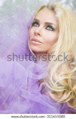 portrait of a beautiful blond woman with long, curly hair that sits behind the glass window, wrapped in a purple tulle