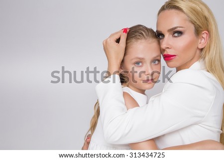 The young girl with the beautiful daughter of an amazing blue eyes and red lips and nails mom blond curly hair long dense sit huddled and dressed in white, a jacket, a dress on a white background