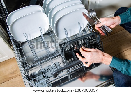 Male hands loading, emptying a dishwasher, taking out a clean fork from a cutlery basket. Household chores with a modern kitchen appliance. An open dishwasher with white plates and silver cutlery.