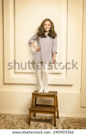 Beautiful happy Lille girl in white room with big windows standing on little wooden stairs and smiling.