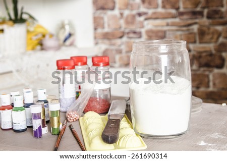 Kitchen table with food dyes in bottles for backing and flour in cup with mixer and bowl of batter on background.