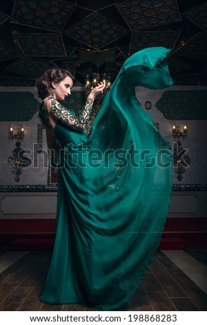 Beautiful woman in a green long dress on a background of richly decorated room. Fashion woman in fluttering dress.