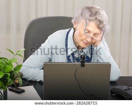 Old teacher is falling asleep in front of laptop screen during lesson on-line. Grey haired old woman is wearing blue blouse and dark blue stone necklace. The glasses will fall off her face soon.