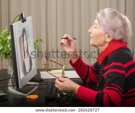 Grey haired expert is teaching student on-line about English pronunciation stress. She is wearing red black jumper. On the screen, woman in headphones is learning how to sound like a native speaker