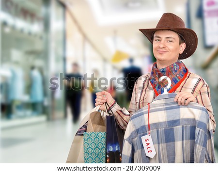 Young cowboy shopping in the mall with shopping bags in left hand. In right hand, he holds blue and white plaid shirt on hangers with tag sale