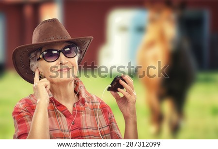 Aged cowgirl in earphones is listening country music with smart phone. She is wearing brown cowboy hat, red plaid shirt and sunglasses. There is a horse on the background out of focus
