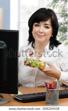 Black haired businesswoman eats healthy vegetable salad for every lunch hour in office. Vertical portrait of middle-aged woman sitting with takeaway transparent container at desk.